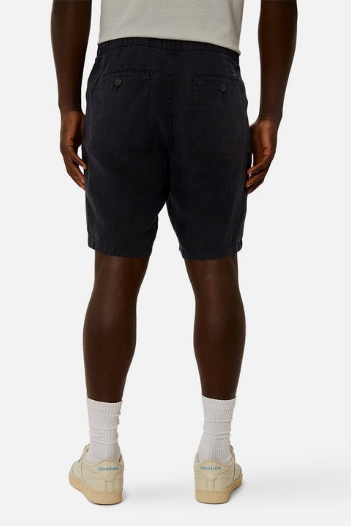 An individual dressed in knee-length Industrie The Baller Linen Shorts featuring a drawstring waist, a light shirt, and white sneakers paired with white socks is standing with their back to the camera against a plain, light-colored background.