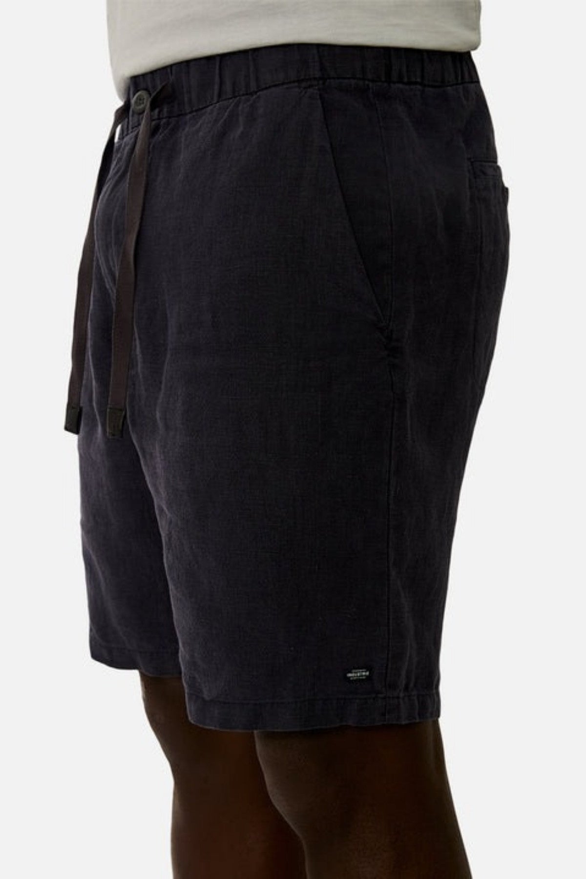 Side view of a person wearing the Industrie The Baller Linen Short, which are knee-length and feature a dark color with an elastic drawstring waist. The fabric has a smooth texture, and there is a small logo at the bottom of the shorts. The background is plain white.