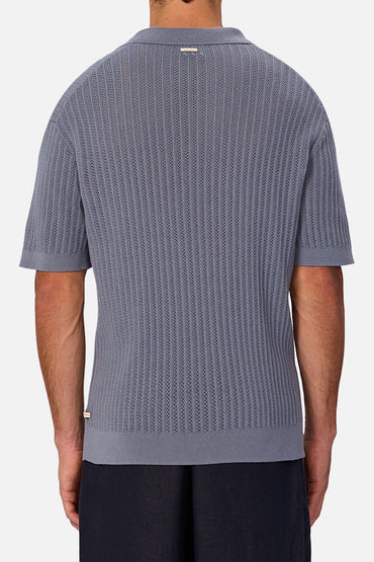 A man is seen from the back wearing a light blue Industrie The Bellano Polo, a men’s 100% cotton knit polo with collar and ribbed hem, paired with dark pants. The background is plain and light-colored.