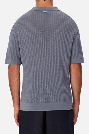 A man is seen from the back wearing a light blue Industrie The Bellano Polo, a men’s 100% cotton knit polo with collar and ribbed hem, paired with dark pants. The background is plain and light-colored.