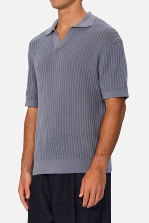 A man wearing the Industrie The Bellano Polo, a light blue 100% cotton knit polo with short sleeves, collar, and open neckline, paired with black pants, stands against a plain light background.