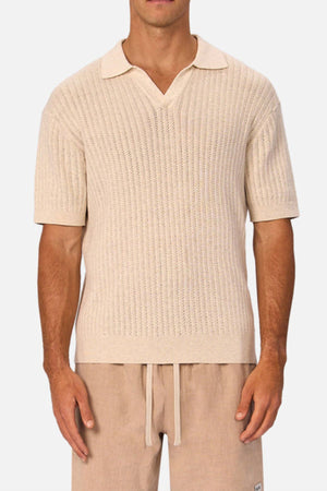 A man wearing the Industrie The Bellano Polo, a light beige short-sleeve knit with a textured pattern and collar, paired with light tan drawstring shorts, stands against a plain white background.