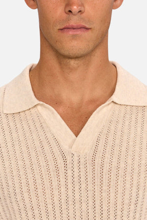A close-up of a person wearing the Industrie The Bellano Polo, a beige men's short-sleeve knit polo with collar and V-neckline. Made from breathable cotton with a vertical perforated pattern. Only the lower half of the face is shown.