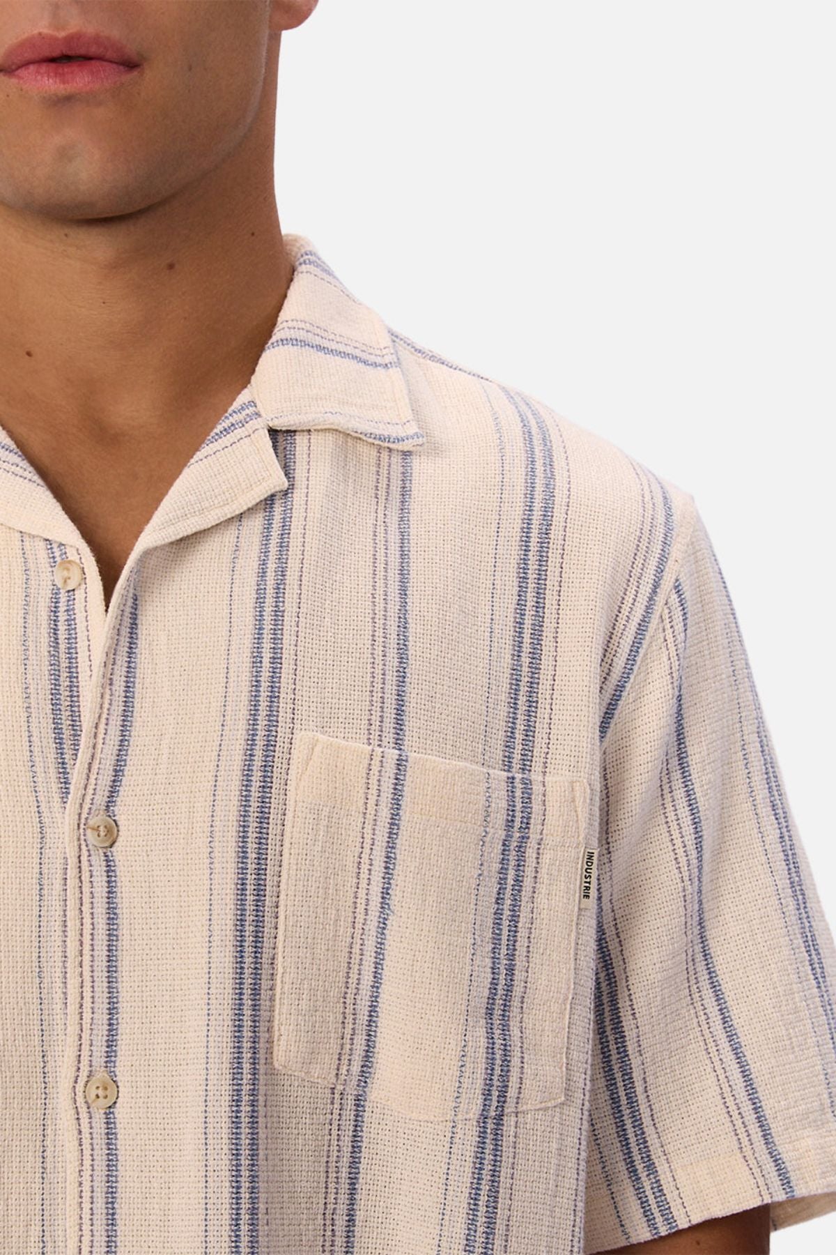 A close-up of a person wearing the Industrie The Bowment Short Sleeve Shirt in cream and blue textured stripes, with a notched collar and chest pocket. Only the lower half of their face and upper chest are visible.