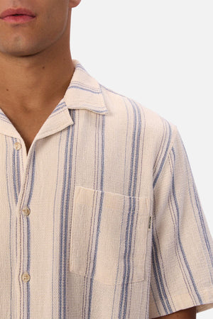 A close-up of a person wearing the Industrie The Bowment Short Sleeve Shirt in cream and blue textured stripes, with a notched collar and chest pocket. Only the lower half of their face and upper chest are visible.