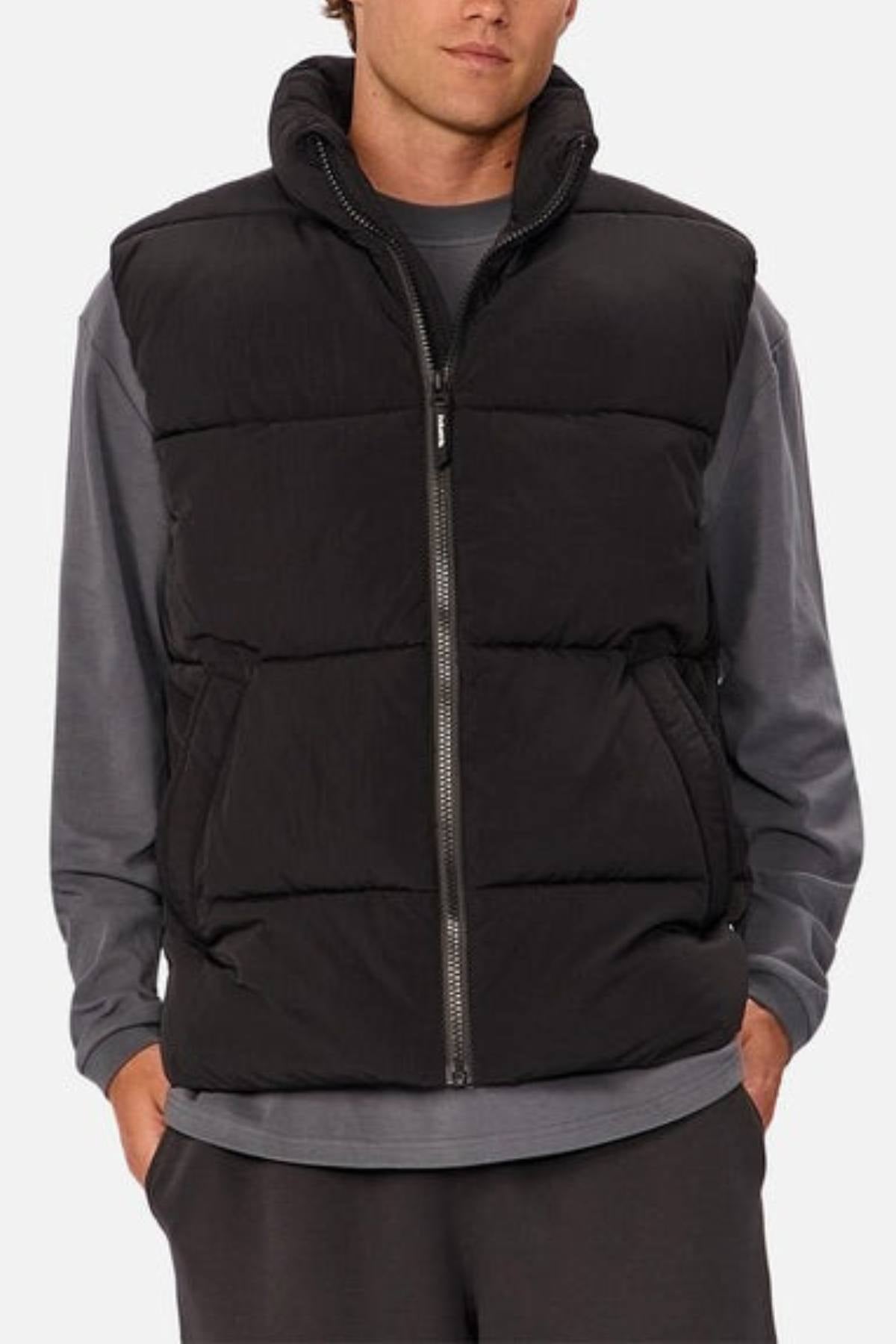 Someone stands with hands in pockets, wearing the Industrie The Busan Puffer Vest—a black nylon vest with Poly-fil padding—over a gray long-sleeve shirt and dark pants, against a plain light background.