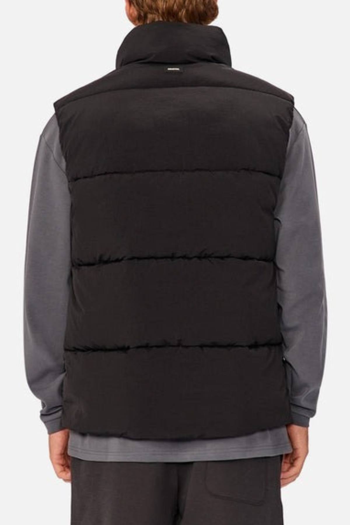 A person is shown from the back wearing the Industrie The Busan Puffer Vest in black with Poly-fil padding over a gray long-sleeve shirt and matching pants. The background is plain and light-colored.