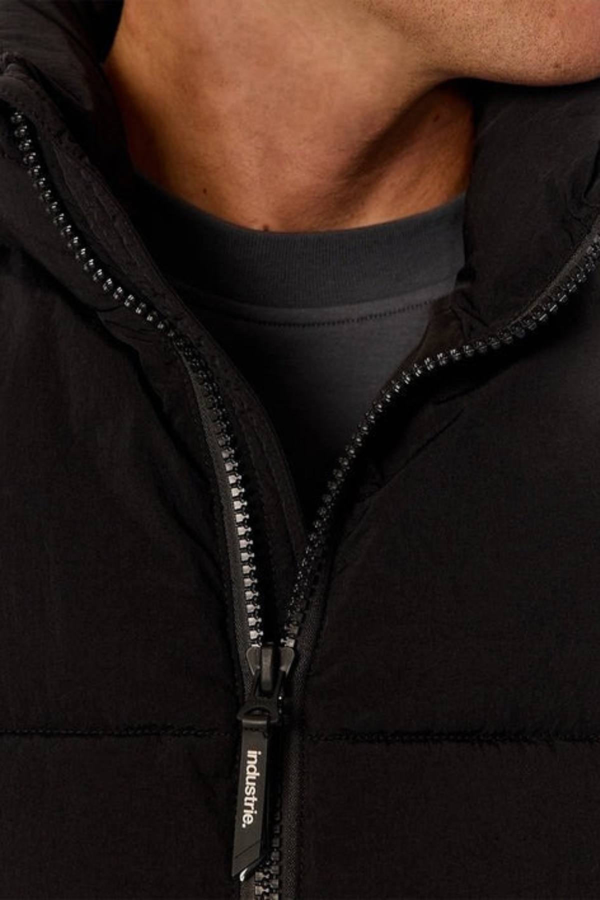 Close-up of a person wearing the Industrie The Busan Puffer Vest in dark nylon, zipper slightly open to reveal a dark crew-neck shirt. Only the lower part of their face and neck are visible.