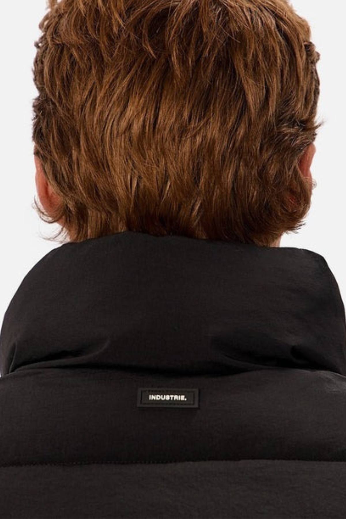 A person with light brown hair is shown from behind, wearing the Industrie The Busan Puffer Vest, a high-collared black nylon vest featuring a small rectangular "INDUSTRIE" label.