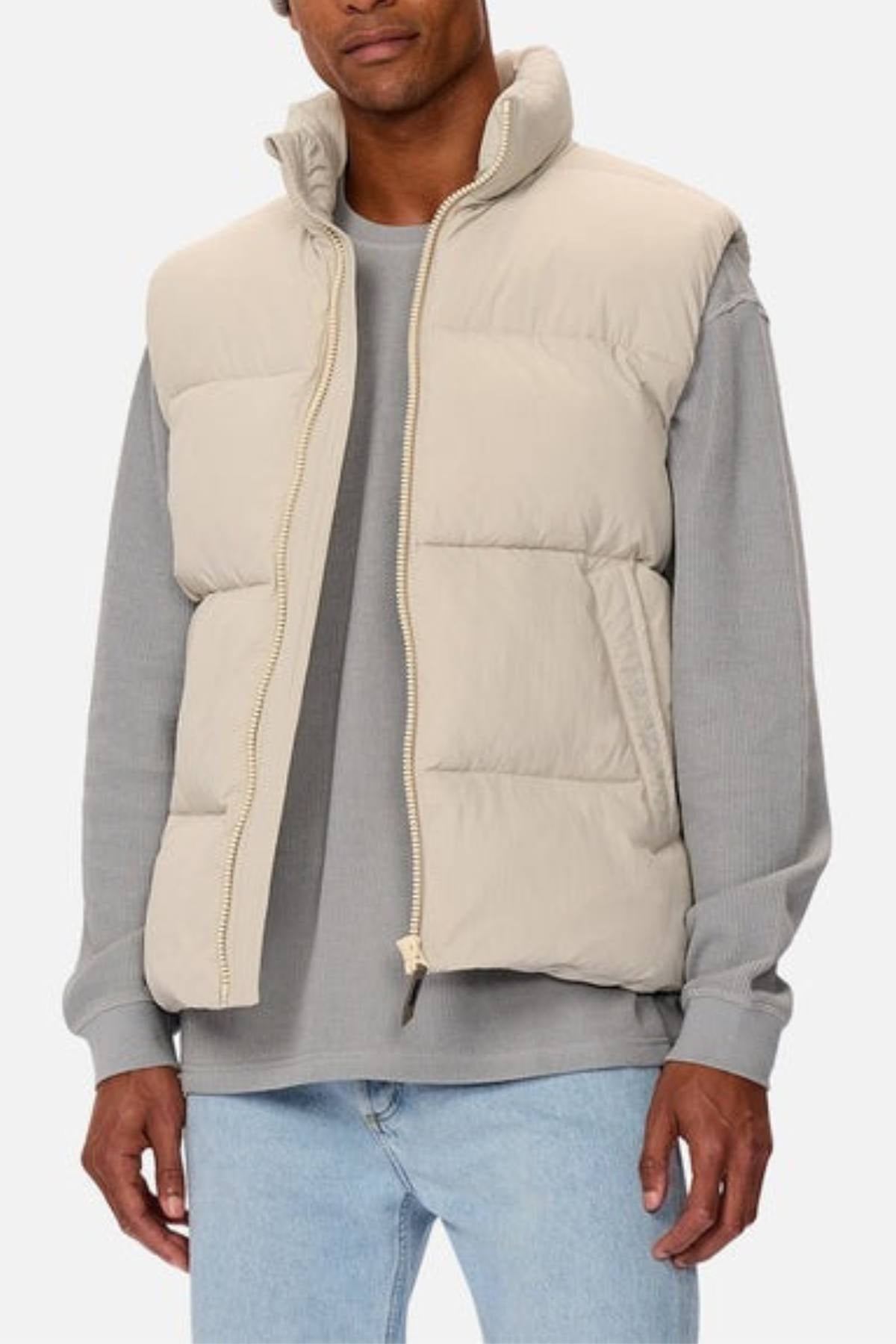 A person wears the Industrie The Busan Puffer Vest in beige nylon with poly-fil padding over a gray long-sleeve shirt and light blue jeans, standing against a plain light background.