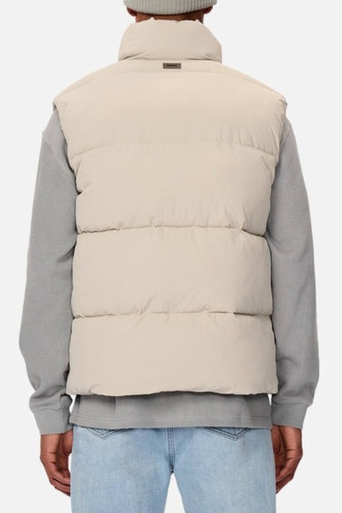 A person seen from behind wears the Industrie The Busan Puffer Vest in beige nylon with poly-fil padding over a gray long-sleeve shirt and light blue jeans, set against a plain light background.