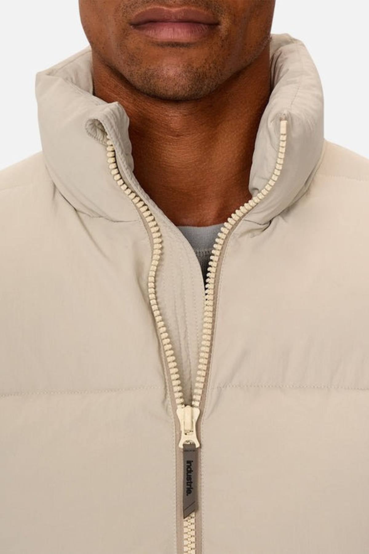 A close-up shows a person wearing the Industrie The Busan Puffer Vest in light beige nylon with Poly-fil padding, a high collar, and partially unzipped front revealing a gray shirt. Only the lower half of their face is visible.