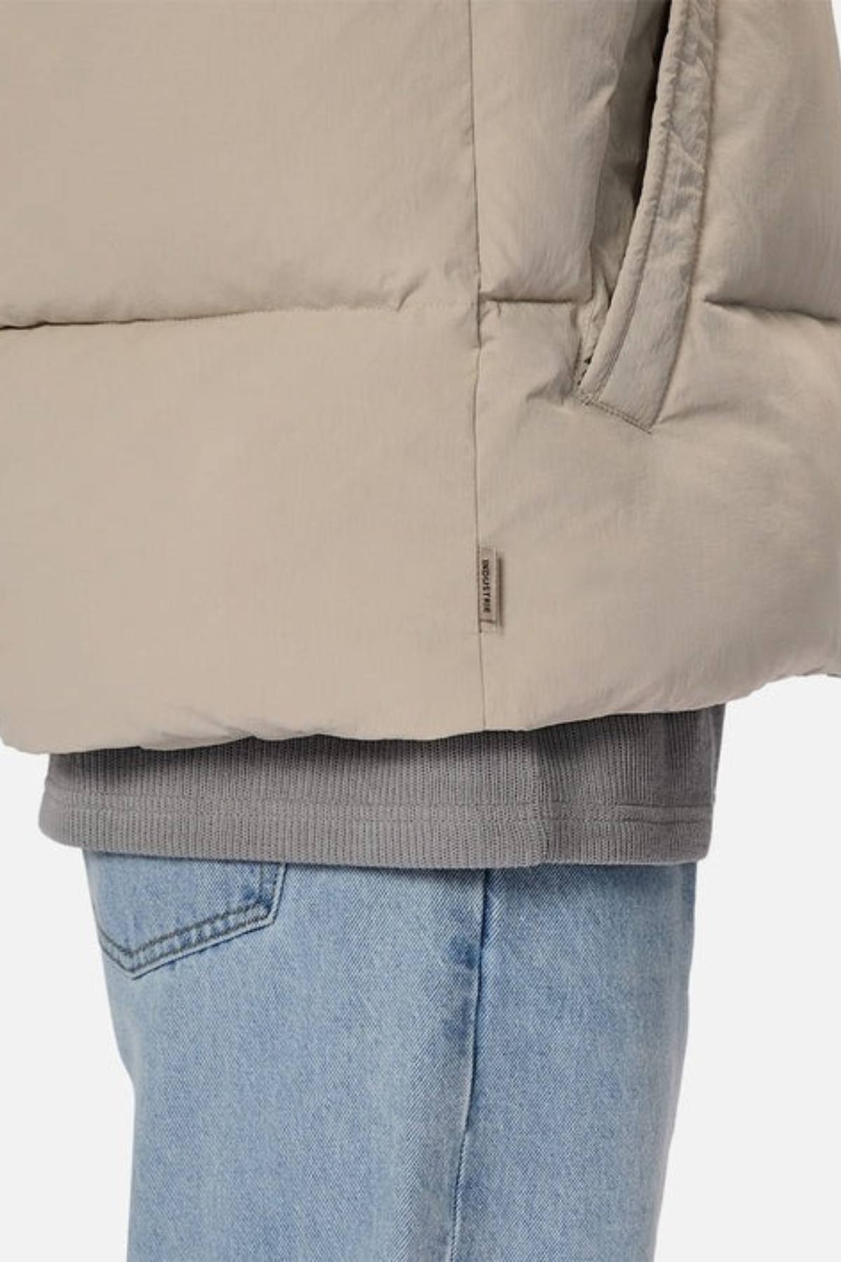 A close-up of a person wearing the Industrie The Busan Puffer Vest in beige nylon with Poly-fil padding over a gray sweatshirt and light blue jeans. Their hand is in the vest pocket against a plain, light background.
