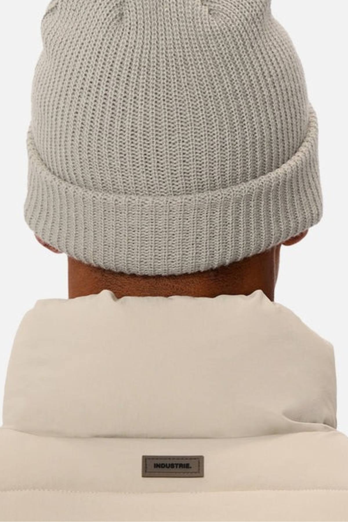 A person is seen from behind wearing a light gray knit beanie and the Industrie The Busan Puffer Vest with Poly-fil Padding. A small rectangular "INDUSTRIE" label appears on the vest's collar.