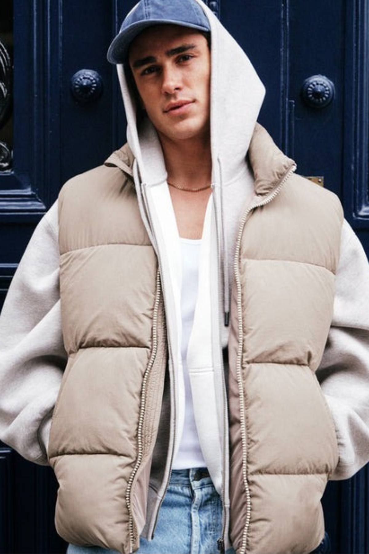 A young man stands before a dark blue door, wearing Industrie The Busan Puffer Vest with Poly-fil padding over a grey hoodie, white tee, blue jeans, and a blue baseball cap, looking confidently at the camera.