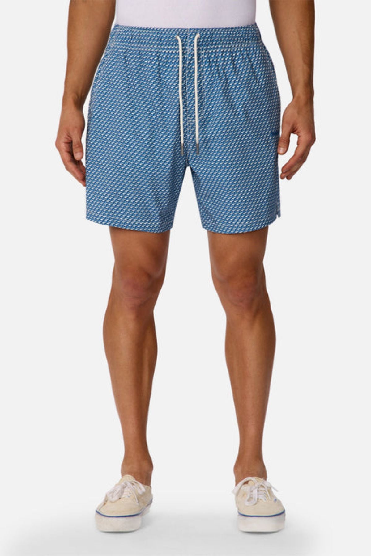 A man wears Industrie The Cayman Swim Short in a blue abstract pattern with a white drawstring, paired with a tucked-in white shirt and beige canvas shoes, standing against a plain white background.