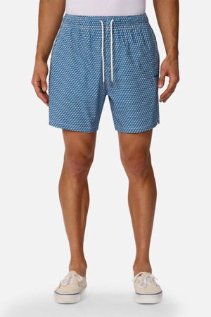 A man wears Industrie The Cayman Swim Short in a blue abstract pattern with a white drawstring, paired with a tucked-in white shirt and beige canvas shoes, standing against a plain white background.