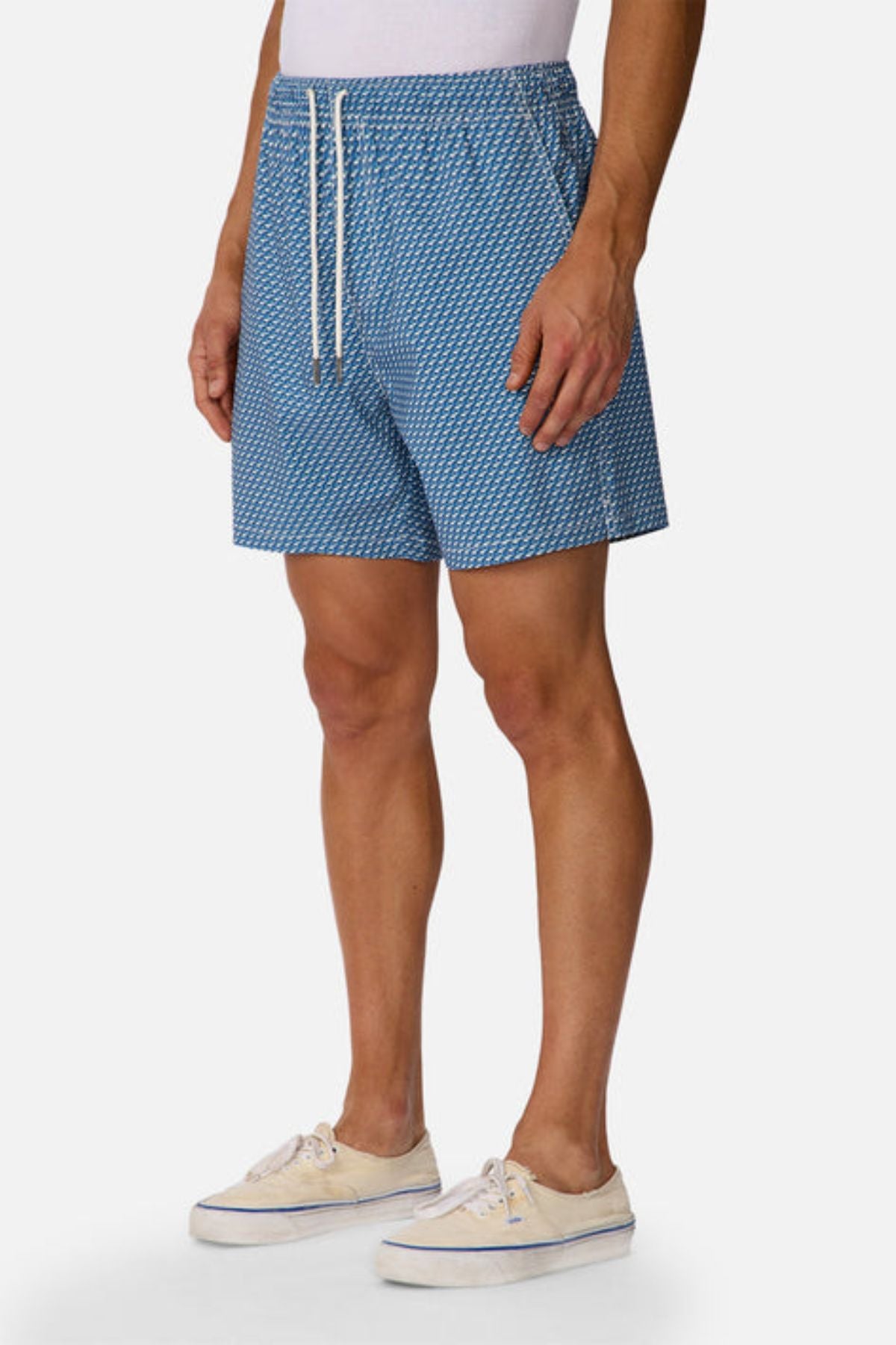 A person wearing Industrie The Cayman Swim Short with an abstract blue pattern, a white shirt, and beige sneakers stands against a plain white background.