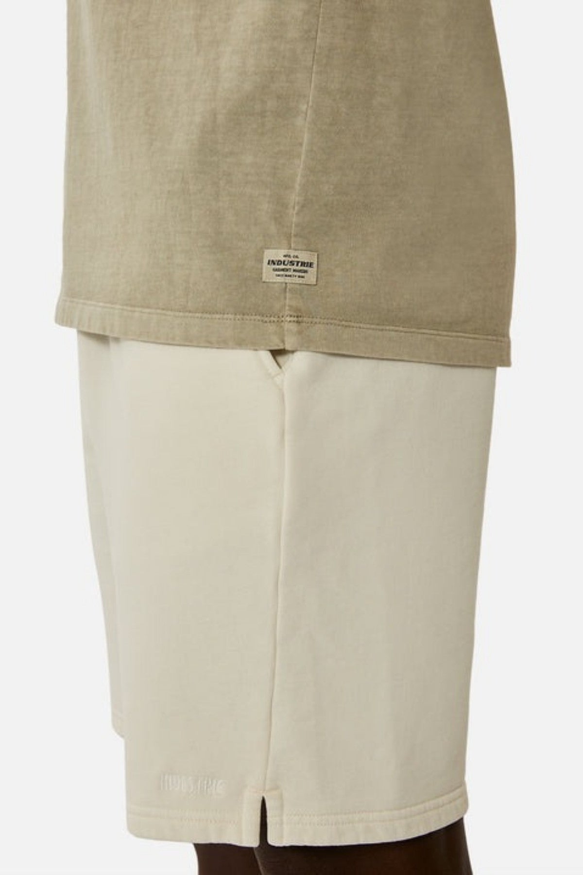 A close-up of a person wearing light beige shorts paired with the Industrie The Del Sur Sleeveless Tee in a khaki, 100% cotton fabric. The shorts exhibit a subtle texture, while the sleeveless tee has an oversized fit and includes a small label near the hem. The background is plain and white.