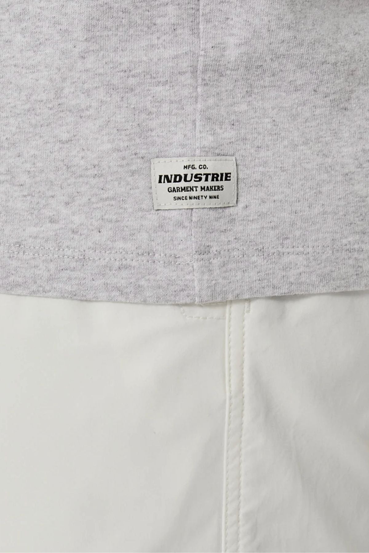 A close-up of the Industrie The Del Sur Sleeveless Tee in light grey cotton, featuring a small white "INDUSTRIE GARMENT MAKERS" label above white pants. The stitching, texture, and oversized fit are clearly shown.