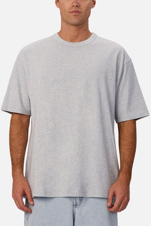 A man wearing an oversized light gray Industrie The Del Sur Tee made from heavyweight fabric and light blue jeans stands facing forward against a white background.