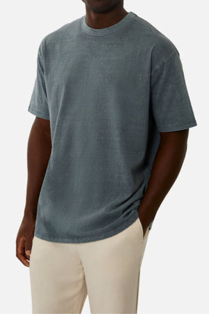 A man in an oversized, faded blue Industrie The Del Sur Tee crafted from heavyweight fabric and light beige pants stands with one hand in his pocket against a plain white background.