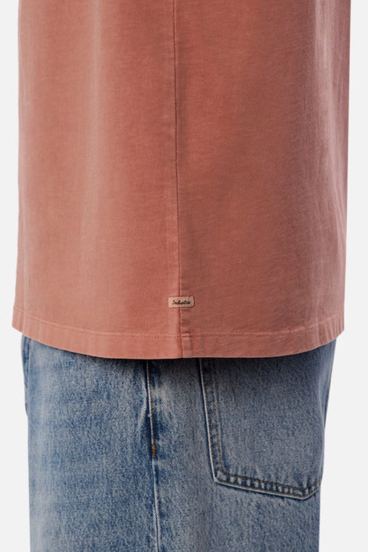 A close-up shows someone wearing the Industrie The Del Sur Tee in a faded pink shade, characterized by its oversized fit and a small tag near the hem. Teamed with light blue jeans, the heavyweight fabric and relaxed design offer an effortlessly casual appearance, drawing attention to the lower half of the tee and jeans.