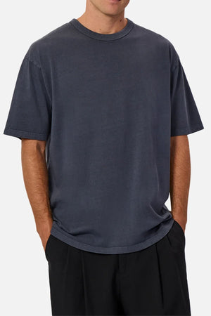 A person wears Industrie The Del Sur Tee, a loose-fit, heavyweight short-sleeve dark gray T-shirt, and black pants with hands in pockets, shown from shoulders to thighs against a plain background.