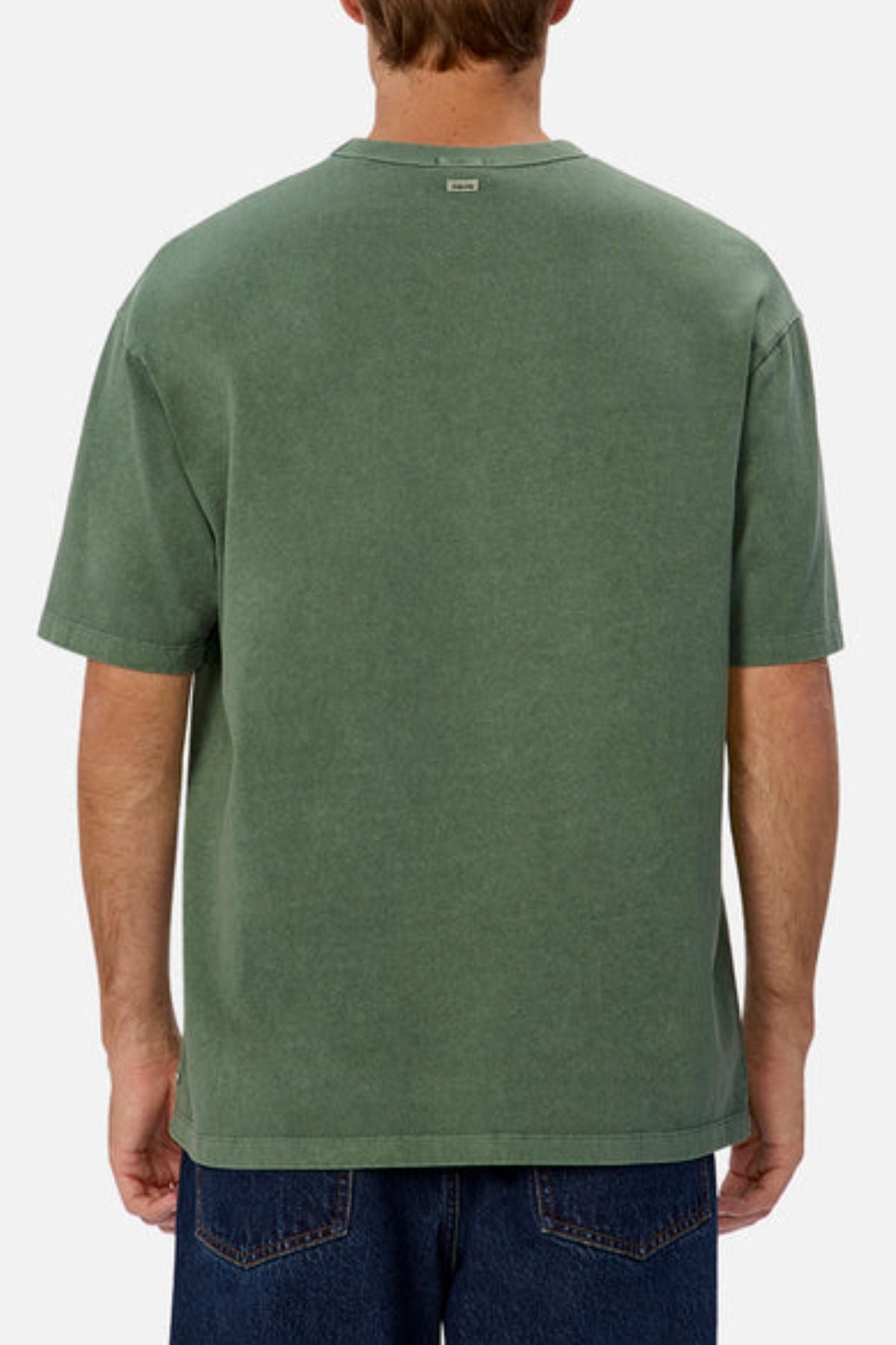 Dressed in Industrie The Del Sur Tee, an oversized fit t-shirt in a faded green shade crafted from heavyweight fabric, and blue jeans, an individual stands with their back to the camera against a plain white background.