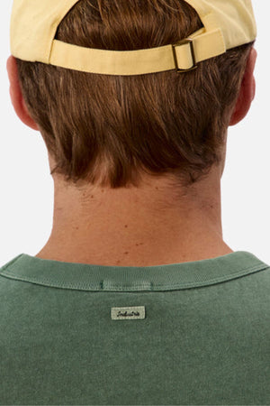Back view of a person wearing a yellow cap and a green Industrie The Del Sur Tee with an oversized fit. The hair is medium-length and brown, and a small tag is visible on the tee's collar. The heavyweight fabric stands out against the plain white background.
