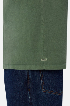 A close-up shot captures someone wearing Industrie The Del Sur Tee in green, matched with blue jeans. The image highlights the hem, where a small label is visible on the left side. This tee offers both comfort and style with its heavyweight fabric and oversized fit.