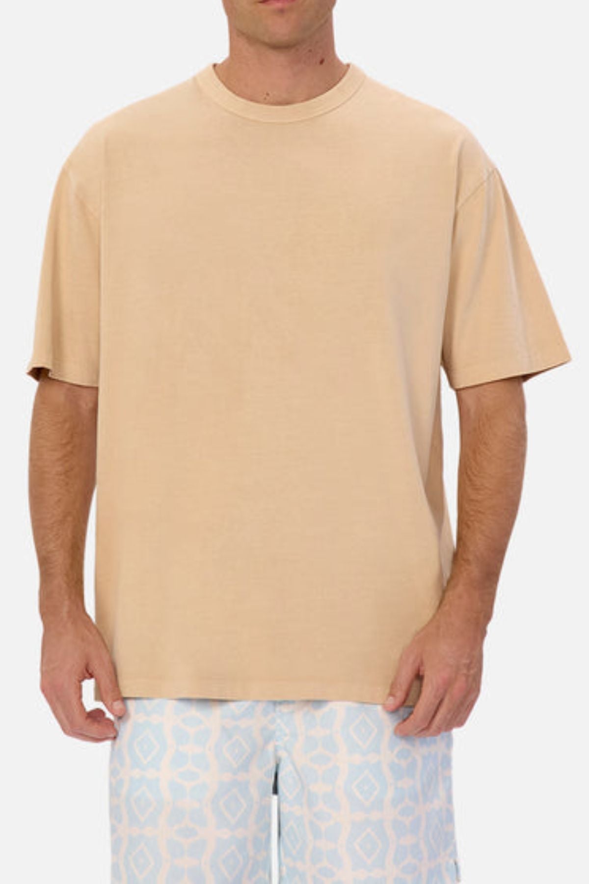 A person wearing an oversized fit beige tee from the Industrie Del Sur collection and patterned blue and white shorts stands against a plain background. The shorts showcase a geometric diamond design, while the heavyweight fabric of the tee complements their relaxed stance with arms by their sides.
