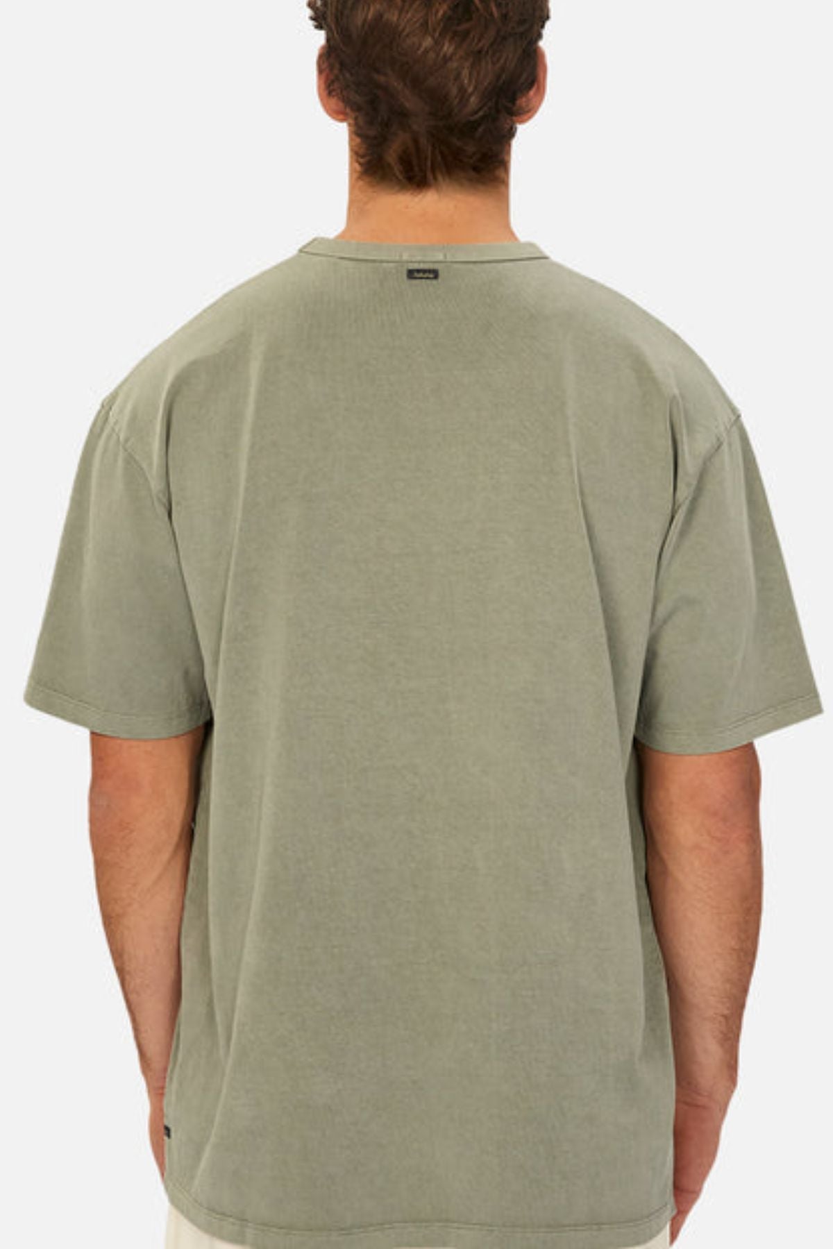 A man with short brown hair is seen from behind wearing the Industrie The Del Sur Tee in heavyweight faded olive green and light shorts, set against a plain white background.