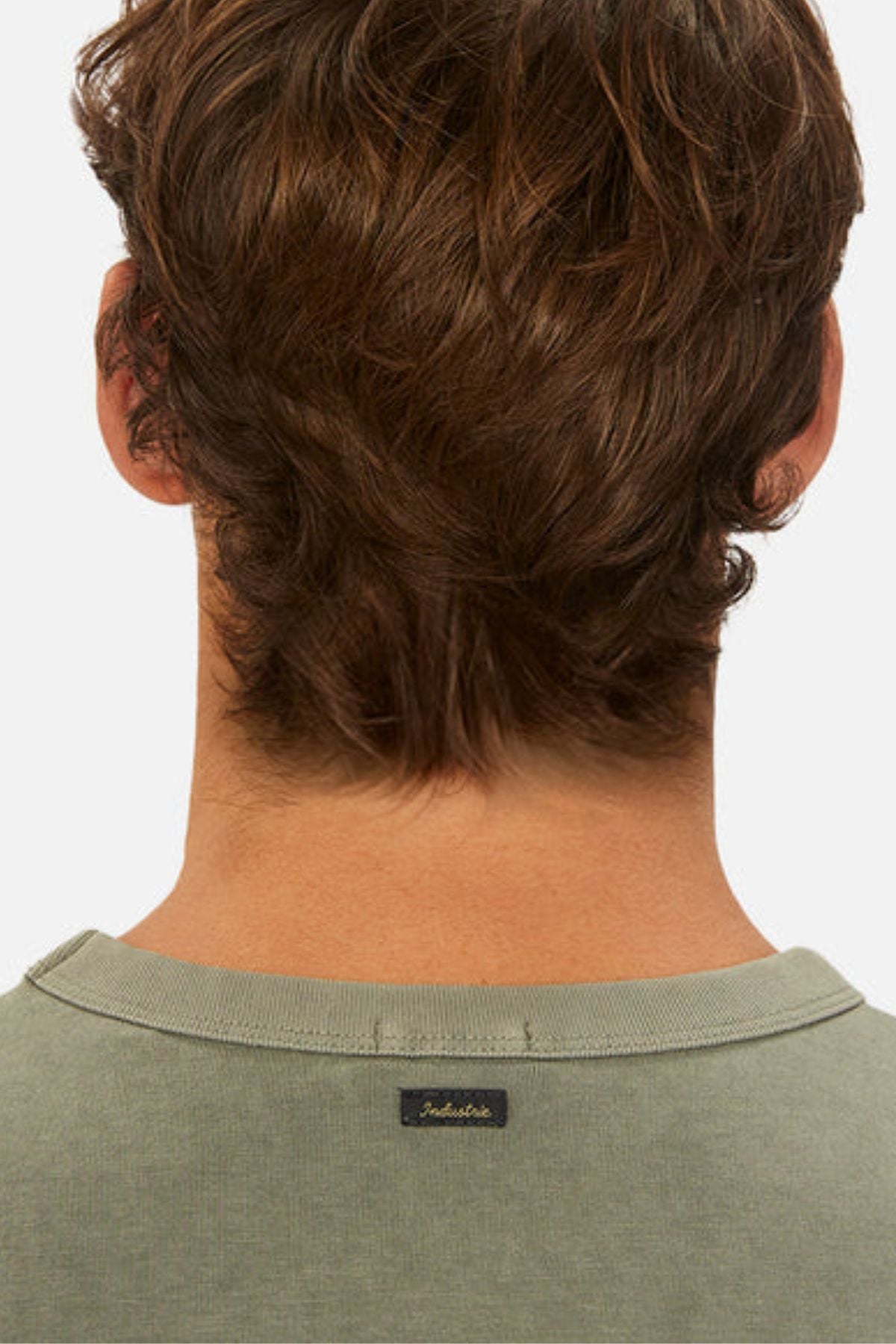 A person with brown, wavy hair is shown from behind in an oversized green Industrie The Del Sur Tee, made of heavyweight fabric. The shirt has a small black Industrie logo on the collar and is set against a plain white background.