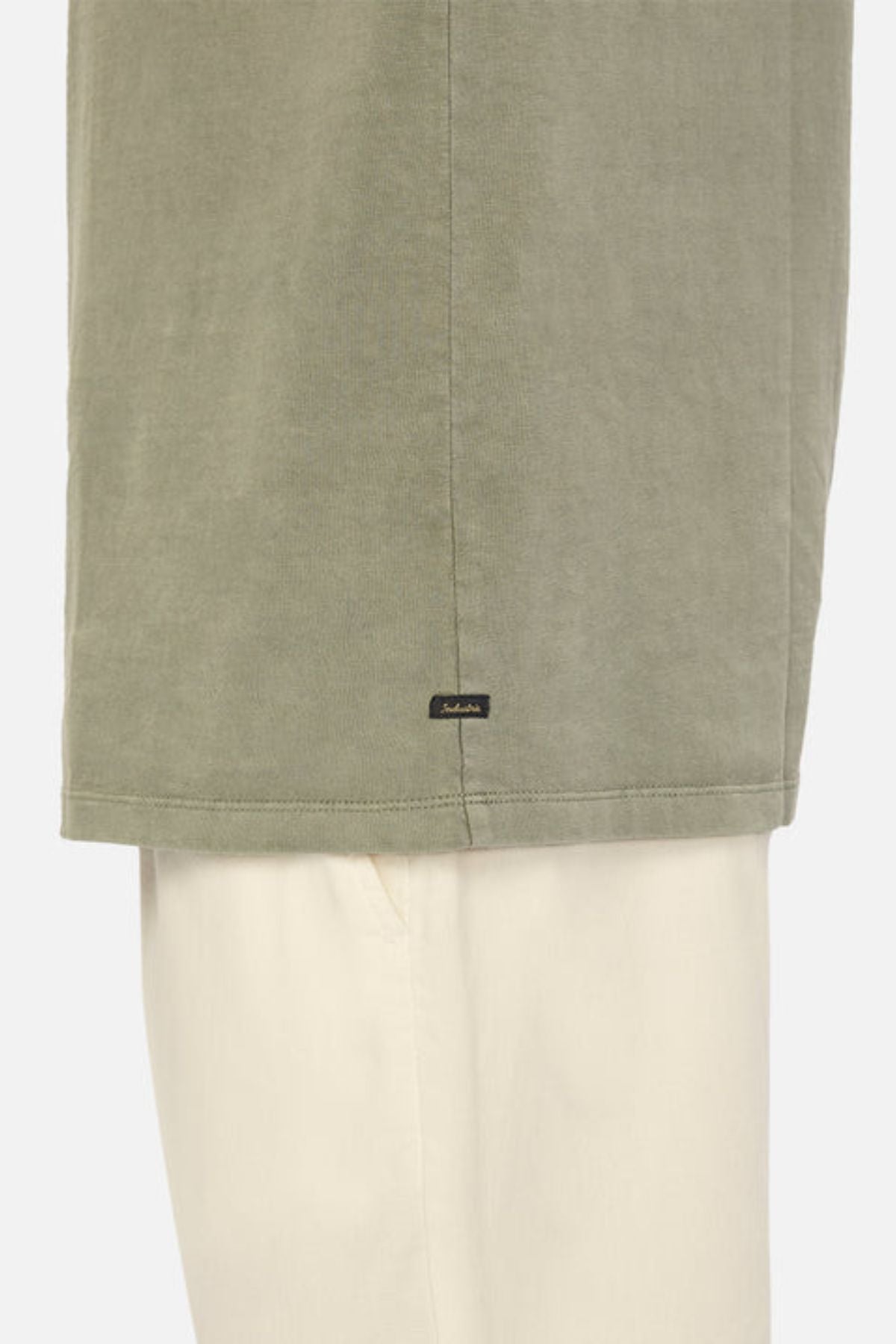 A close-up shows the lower part of the Industrie The Del Sur Tee in khaki green heavyweight fabric, with a small black logo tag above the hem, styled over cream-colored pants.