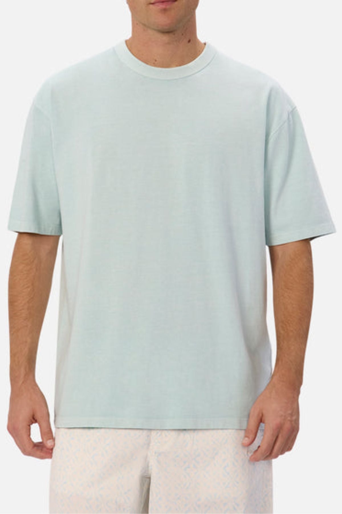 A man is dressed in an Industrie The Del Sur Tee in light blue, featuring an oversized fit, paired with light-colored shorts adorned with a subtle pattern. The tee's heavyweight fabric contrasts against the plain white background as he stands with his arms by his sides.