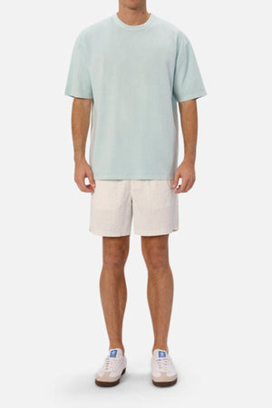 A person wearing an oversized, light green Industrie The Del Sur Tee featuring heavyweight fabric stands against a plain white background. They have paired it with beige shorts adorned with small patterns and completed the look with white sneakers. Their hands rest relaxed by their sides.