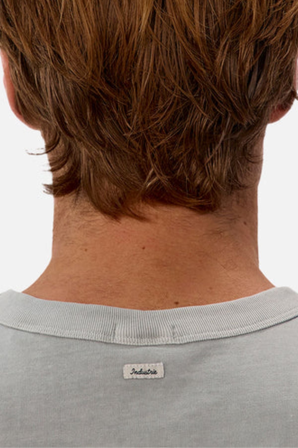 Close-up of the back of a person's neck and head with short, light brown hair, wearing an oversized light gray Industrie The Del Sur Tee featuring a small white "Industrie" label near the collar.
