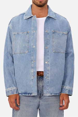 A person wears the Industrie The Denim Overshirt in a relaxed fit light blue wash over a white shirt and blue jeans. Featuring two chest pockets and metal buttons, it’s worn open for a classic vintage style against a plain, light background.