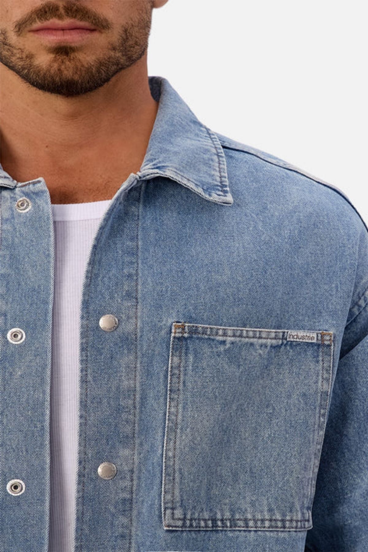 A close-up of a man in the Industrie The Denim Overshirt in light blue, relaxed fit, styled over a white ribbed shirt. The focus is on the jacket’s front pocket and collar, with only the lower half of his face visible.
