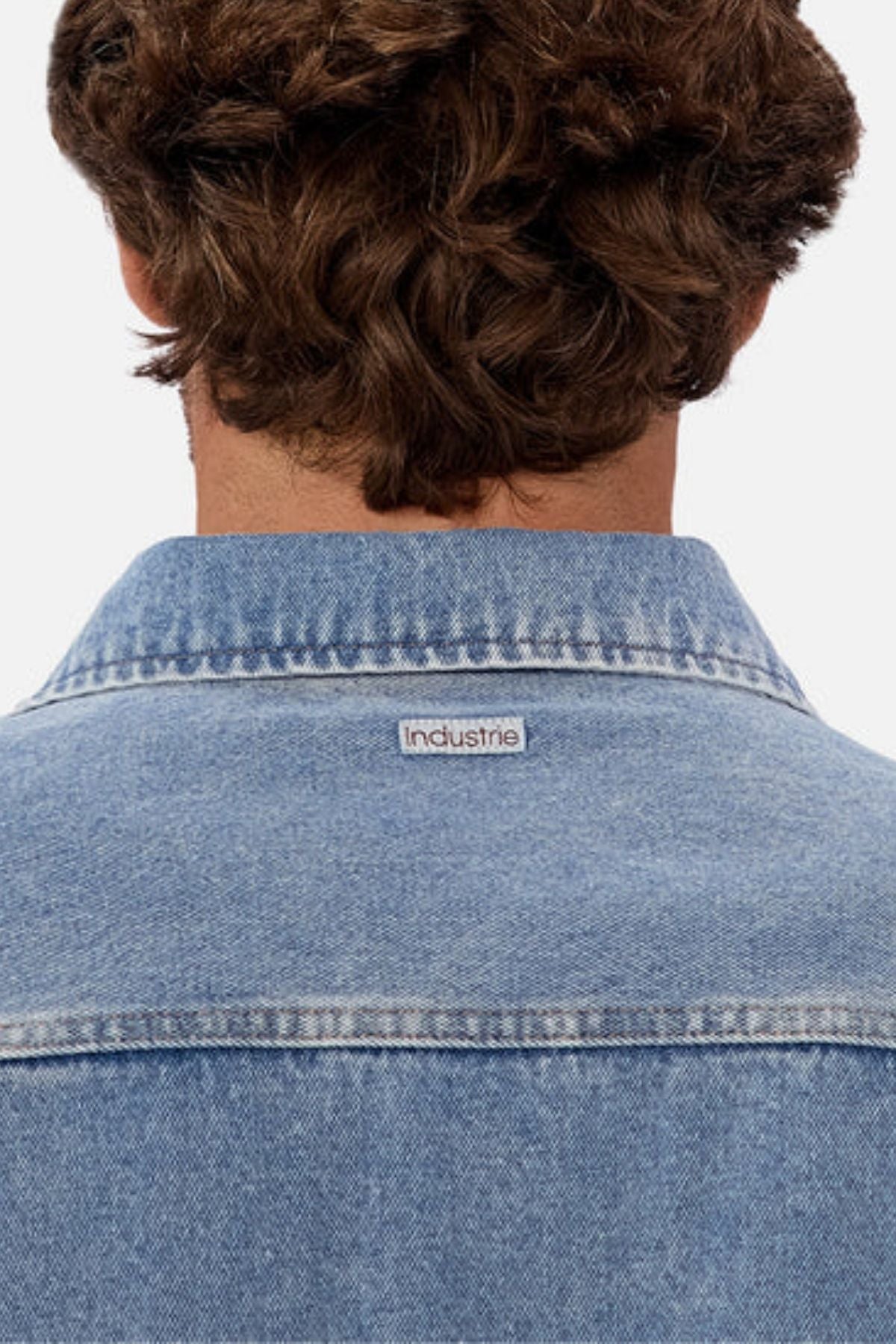 A person with curly brown hair is shown from behind wearing the Industrie The Denim Overshirt in light blue, featuring a small "Industrie" label on the collar, with a relaxed fit and vintage-inspired style.