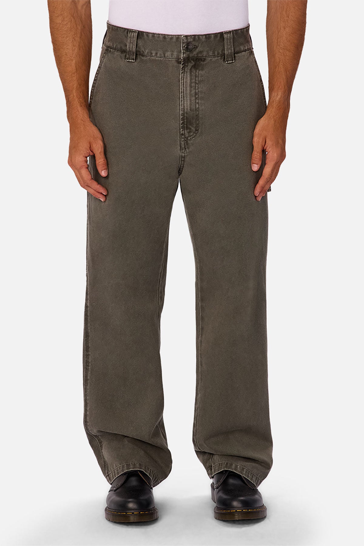 A person wears loose-fitting, faded olive green Industrie The Detroit Carpenter Pant in 100% cotton canvas, a tucked white shirt, and black shoes, standing against a white background for a classic street style look.