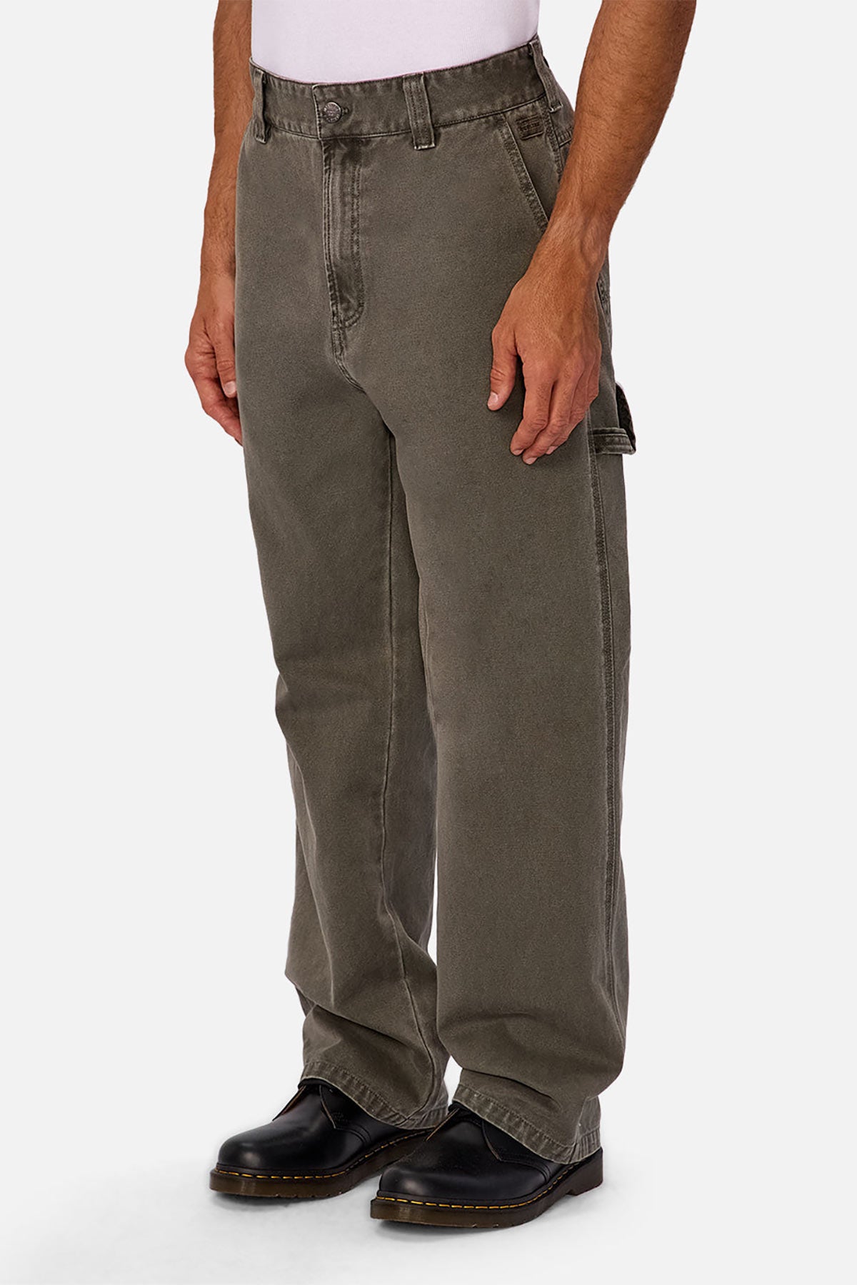 Someone wearing loose-fitting, faded olive-green Industrie The Detroit Carpenter Pant in 100% cotton canvas, a white shirt, and black leather boots stands with arms relaxed at sides against a plain white background for a street style look.