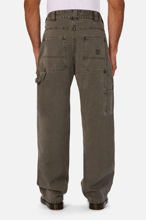 A man stands facing away, wearing Industrie The Detroit Carpenter Pant in olive green 100% cotton canvas with large back pockets, a side hammer loop, and brown shoes—showcasing street style with visible stitching and a casual, worn-in look.