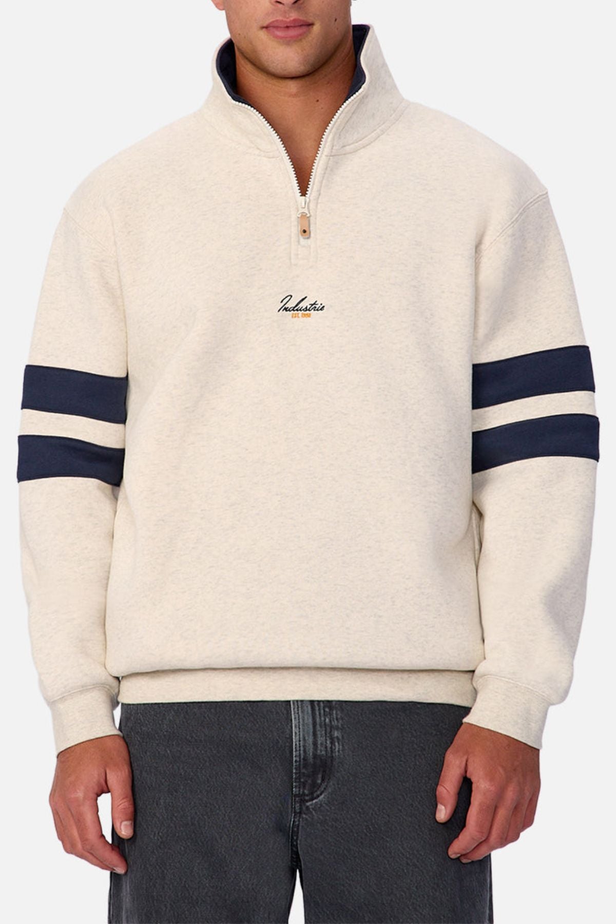 A man wears the Industrie The East Village Track Top in light beige with dark blue double sleeve stripes and small chest embroidery, paired with dark gray jeans. The background is plain white.
