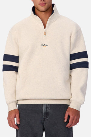 A man wears the Industrie The East Village Track Top in light beige with dark blue double sleeve stripes and small chest embroidery, paired with dark gray jeans. The background is plain white.