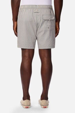 A person is shown from the back, standing against a plain white background, wearing Industrie The Formosa Swim Short in a blue and white striped pattern paired with light-colored slip-on shoes.