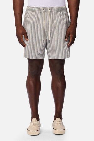 A person wearing a white tank top, beige sneakers, and the Industrie The Formosa Swim Short—featuring light blue and beige stripes with a drawstring—stands facing forward against a plain background.