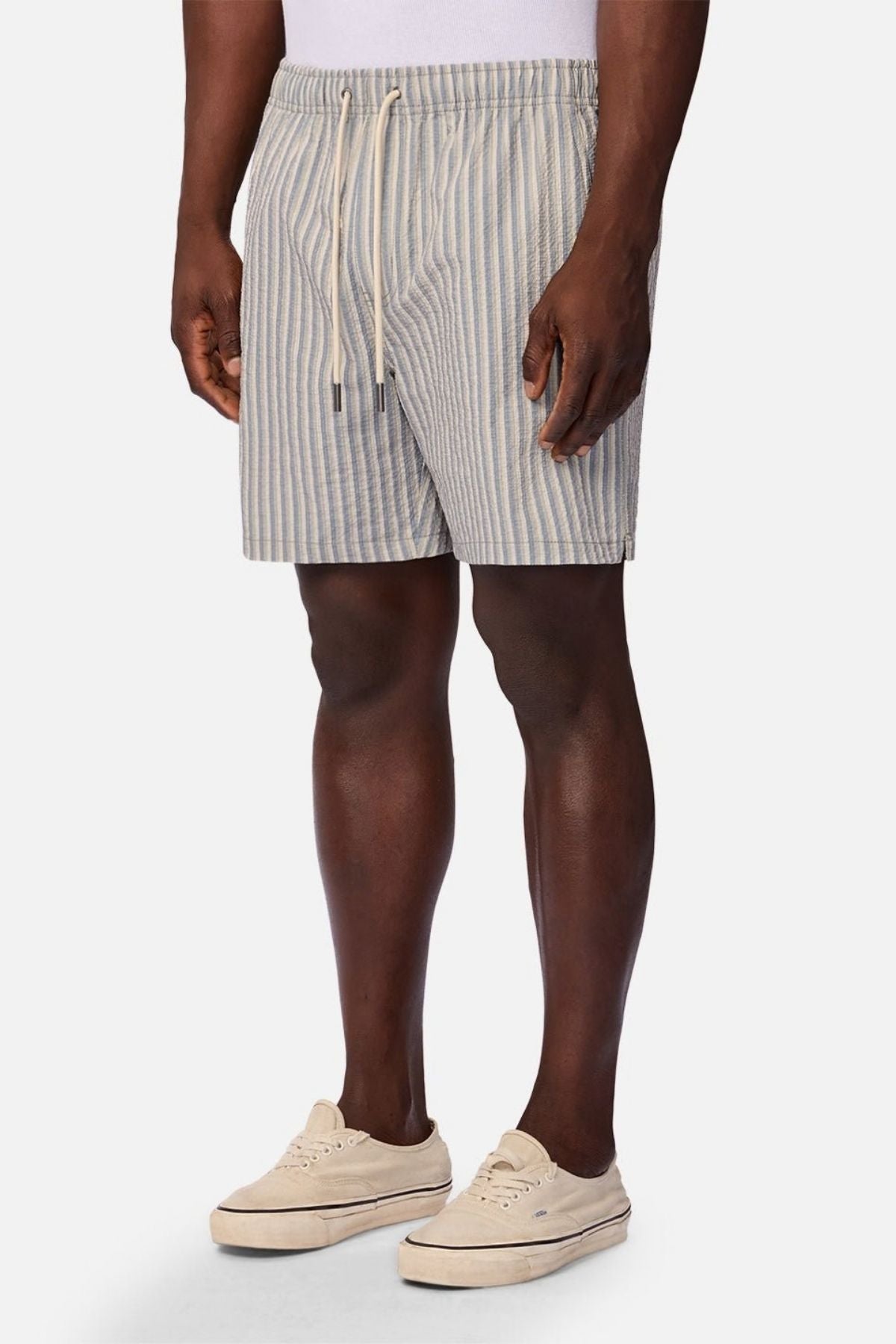 Wearing the Industrie The Formosa Swim Short in light blue and white stripes, a person stands against a plain background paired with a white shirt and beige sneakers.