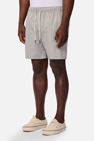 Wearing the Industrie The Formosa Swim Short in light blue and white stripes, a person stands against a plain background paired with a white shirt and beige sneakers.